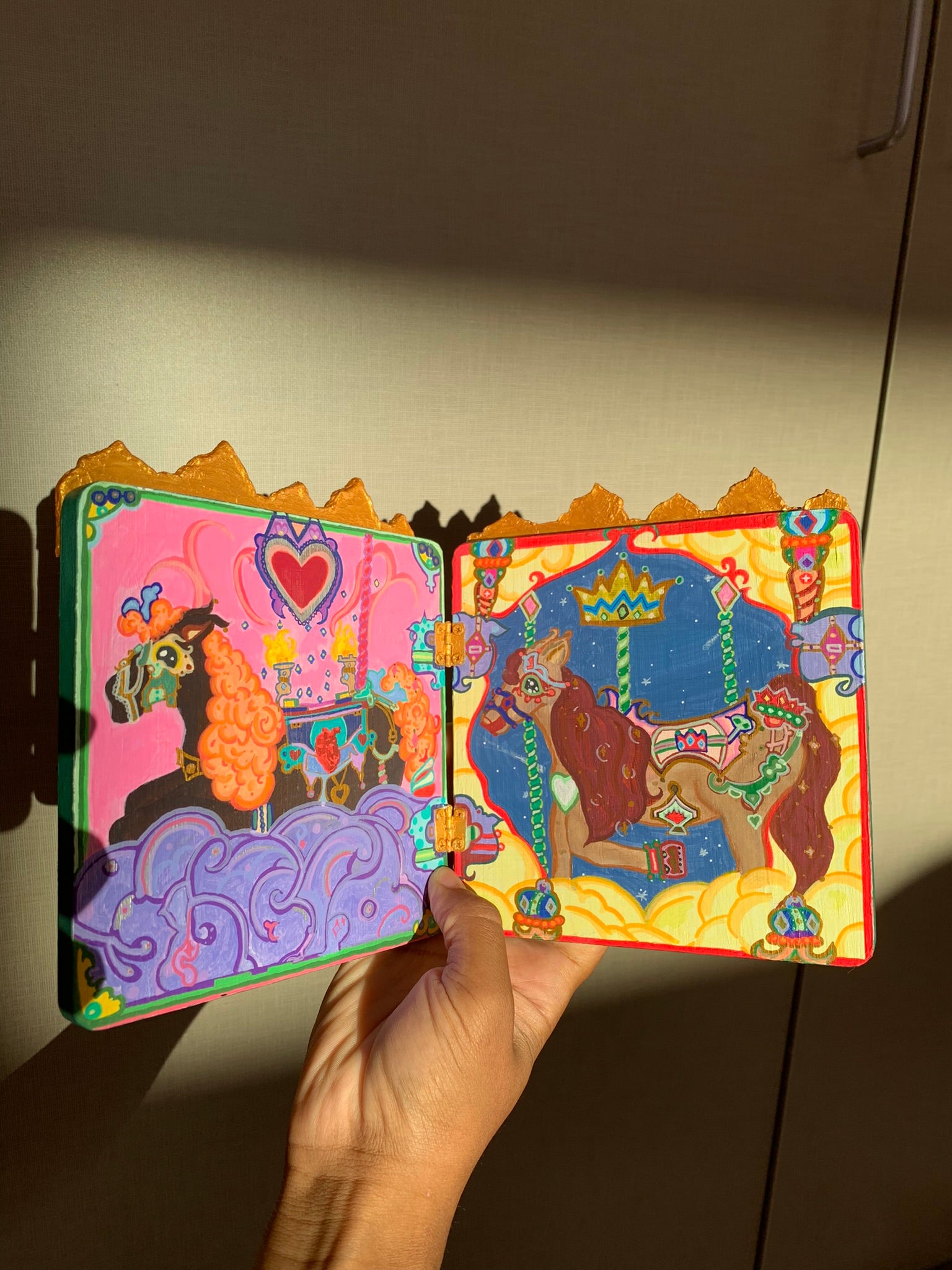 Colorful carousel-themed pony mini painting artwork. The Miraculous Carousel Girls wooden painting book, vibrantart, equineart, carouselride, merrygoroundmagic, immersiveart, nostalgickidcore, hopefulart, innocentjoy, nurserydecor, babygirlroom, childlikemagic, playfulimagination, joyfulplaytime, majesticlookbook, nurserybabyart, babygirldecor, cherishablekeepsakes, functionalsurrealism, surrealismsculpture
Colorful children's book with animal illustrations held by a hand against a neutral background