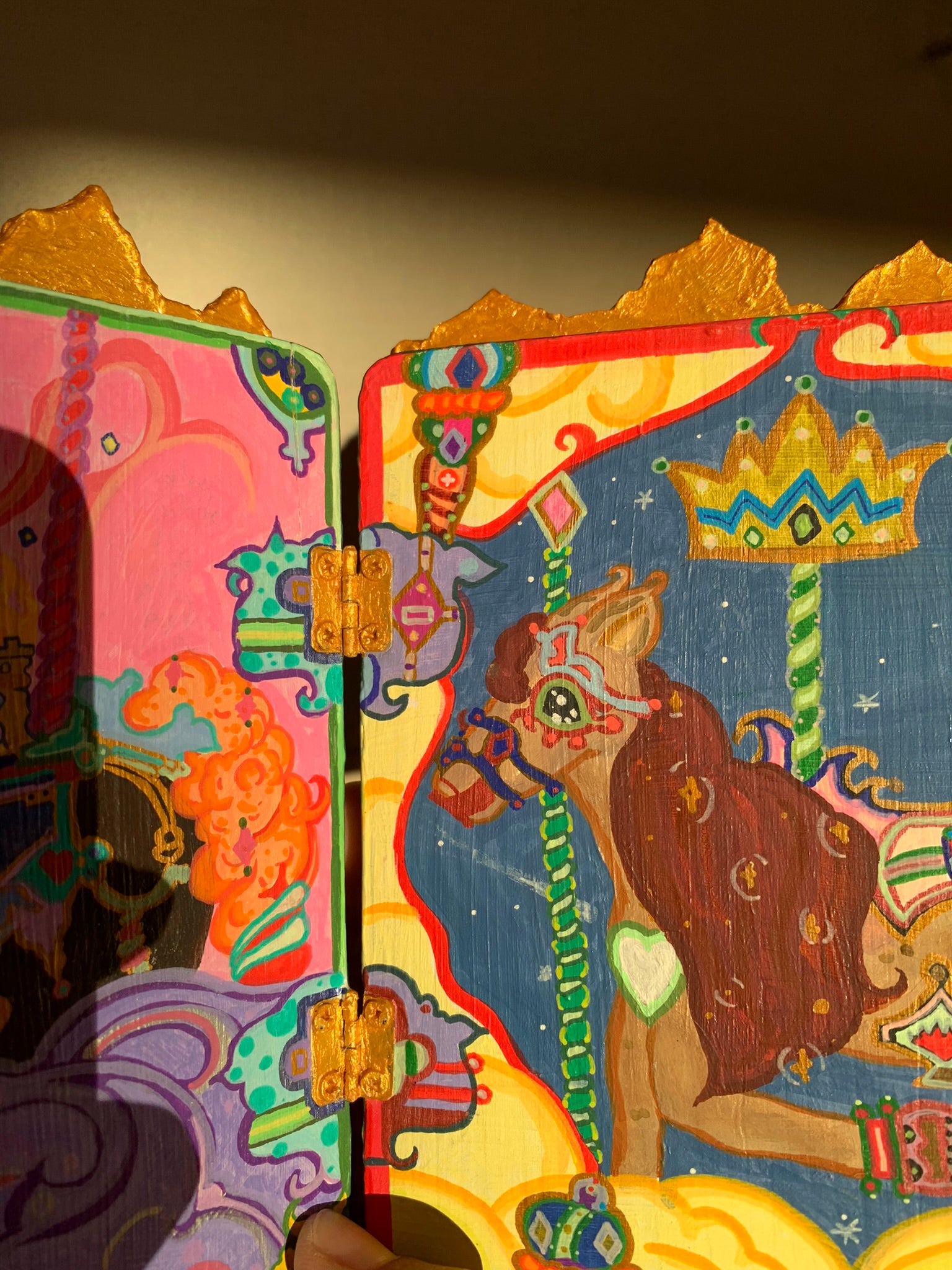 Colorful wooden box with intricate designs and a horse illustration on a dark background. Colorful carousel-themed artwork on a small easel. The Miraculous Carousel Girls wooden painting book, vibrantart, equineart, carouselride, merrygoroundmagic, immersiveart, nostalgickidcore, hopefulart, innocentjoy, nurserydecor, babygirlroom, childlikemagic, playfulimagination, joyfulplaytime, majesticlookbook, nurserybabyart, babygirldecor