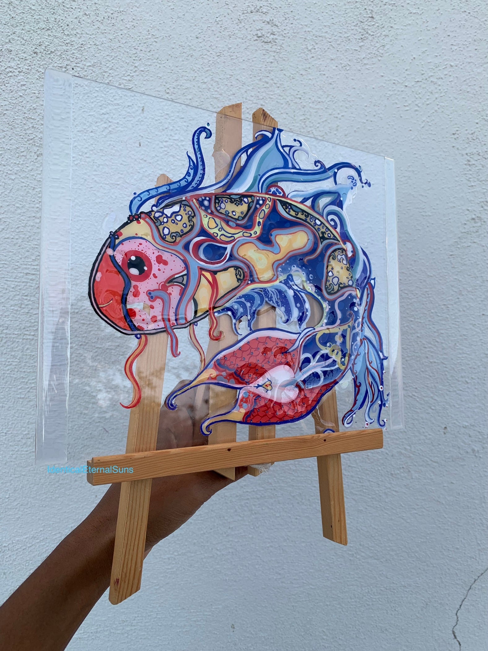 Colorful abstract fish drawing on a clear sheet held by a person with a wooden easel against a light gray background. Identicaleternalsuns, art collection, art buyer, Identicaleternalsuns, DenleyTwins, Whimsical, radiant, otherworldly, glorious, fantastical, eternal, divine, Dreamy Fish, Aquarium wonderland, Frutiger Aero vibes, identicaleternalsuns, Ecstatic, frutiger aero art, fish painting on glass, koi, koifish art, mixed media , fishart, fish glass painting, iridescentart, underwaterart, traditionalart