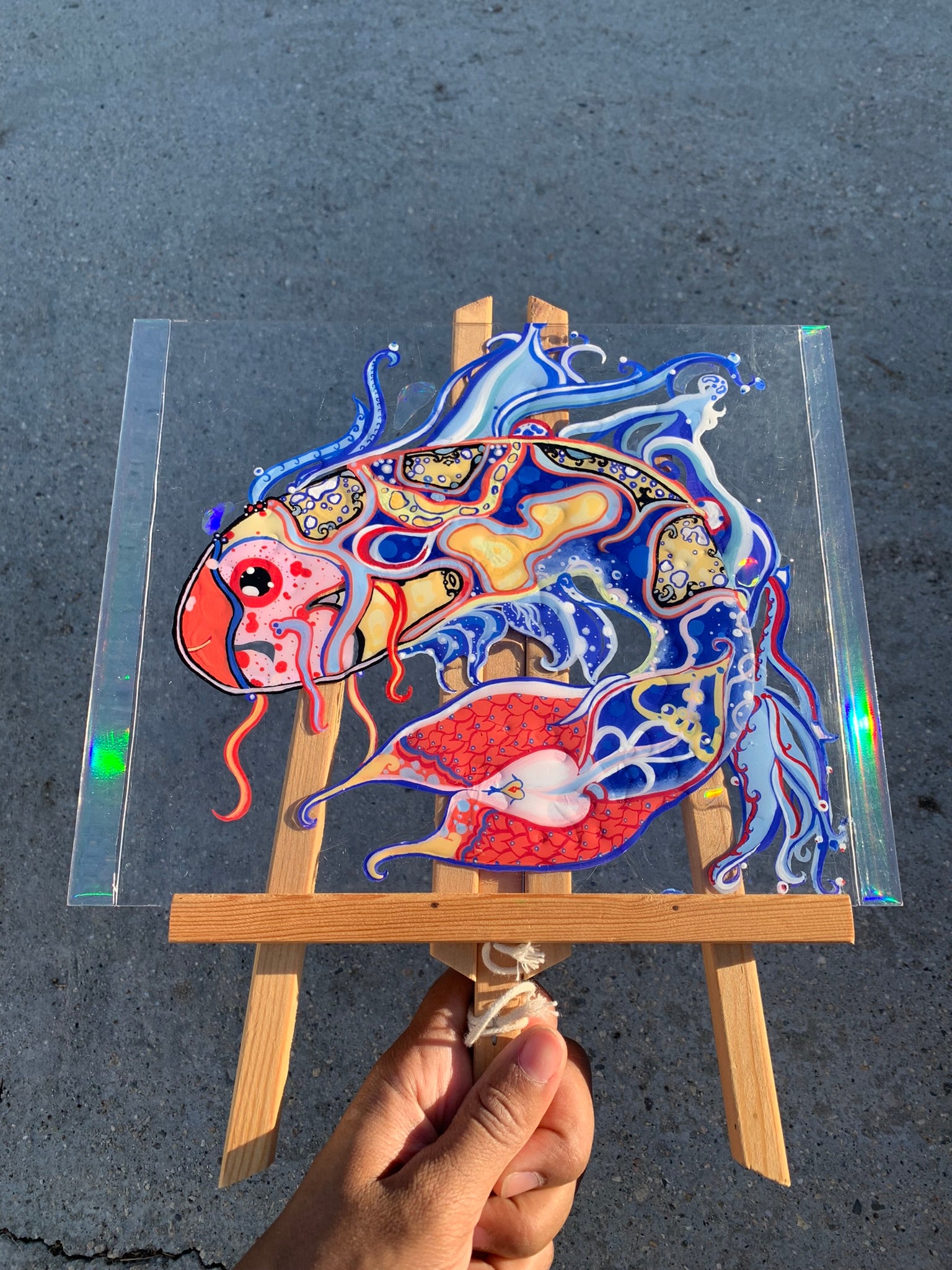 Colorful fish illustration on a clear sheet held by a hand with a black background. Identicaleternalsuns, art collection, art buyer, Identicaleternalsuns, DenleyTwins, Whimsical, radiant, otherworldly, glorious, fantastical, eternal, divine, Dreamy Fish, Aquarium wonderland, Frutiger Aero vibes, identicaleternalsuns, Ecstatic, frutiger aero art, fish painting on glass, koi, koifish art, mixed media , fishart, fish glass painting, iridescentart, underwaterart, traditional art, contemporary art, asian art