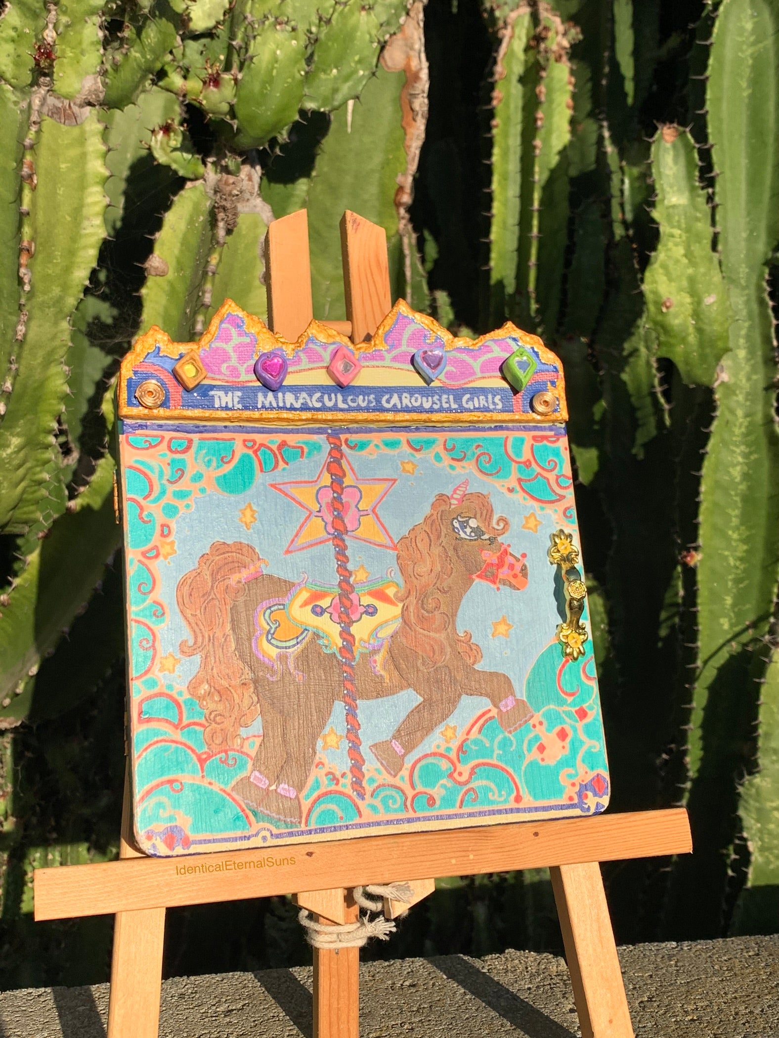Colorful carousel horse painting on an easel with cacti in the background. Colorful carousel-themed pony mini painting artwork. The Miraculous Carousel Girls wooden painting book, vibrantart, equineart, carouselride, merrygoroundmagic, immersiveart, nostalgickidcore, hopefulart, innocentjoy, nurserydecor, babygirlroom, childlikemagic, playfulimagination, joyfulplaytime, majesticlookbook, nurserybabyart, babygirldecor, cherishablekeepsakes, functionalsurrealism, surrealismsculpture