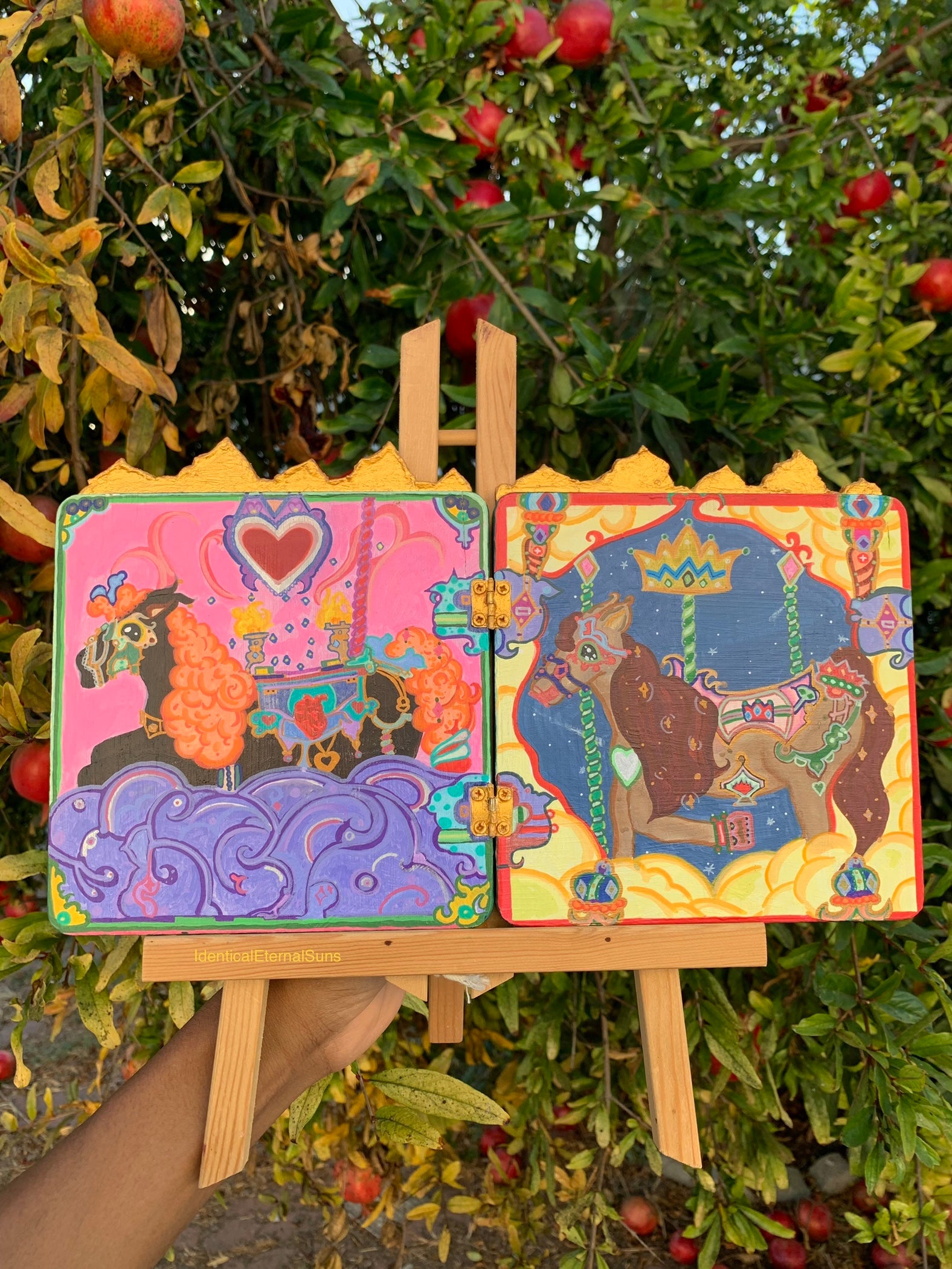 Colorful children's book on an easel with a tree and apples in the background. Colorful carousel-themed artwork on a small easel. The Miraculous Carousel Girls wooden painting book, vibrantart, equineart, carouselride, merrygoroundmagic, immersiveart, nostalgickidcore, hopefulart, innocentjoy, nurserydecor, babygirlroom, childlikemagic, playfulimagination, joyfulplaytime, majesticlookbook, nurserybabyart, babygirldecor, cherishablekeepsakes, functionalsurrealism, surrealismsculpture