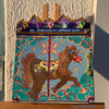 Colorful carousel-themed artwork on a small easel against a textured wall. The Miraculous Carousel Girls wooden painting book, vibrantart, equineart, carouselride, merrygoroundmagic, immersiveart, nostalgickidcore, hopefulart, innocentjoy, nurserydecor, babygirlroom, childlikemagic, playfulimagination, joyfulplaytime, majesticlookbook, nurserybabyart, babygirldecor, cherishablekeepsakes, functionalsurrealism, surrealismsculpture