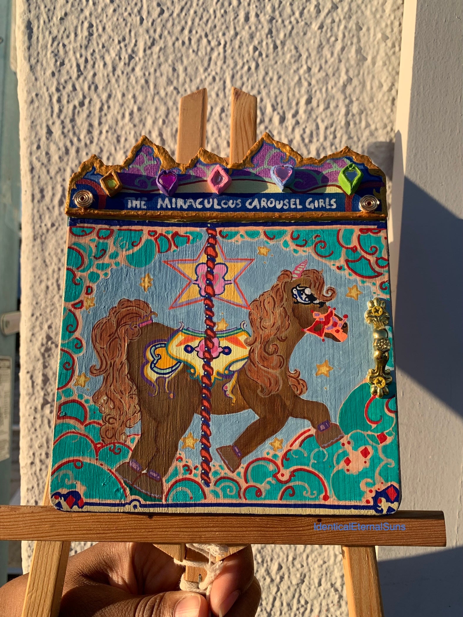Colorful carousel-themed artwork on a small easel against a textured wall. The Miraculous Carousel Girls wooden painting book, vibrantart, equineart, carouselride, merrygoroundmagic, immersiveart, nostalgickidcore, hopefulart, innocentjoy, nurserydecor, babygirlroom, childlikemagic, playfulimagination, joyfulplaytime, majesticlookbook, nurserybabyart, babygirldecor, cherishablekeepsakes, functionalsurrealism, surrealismsculpture