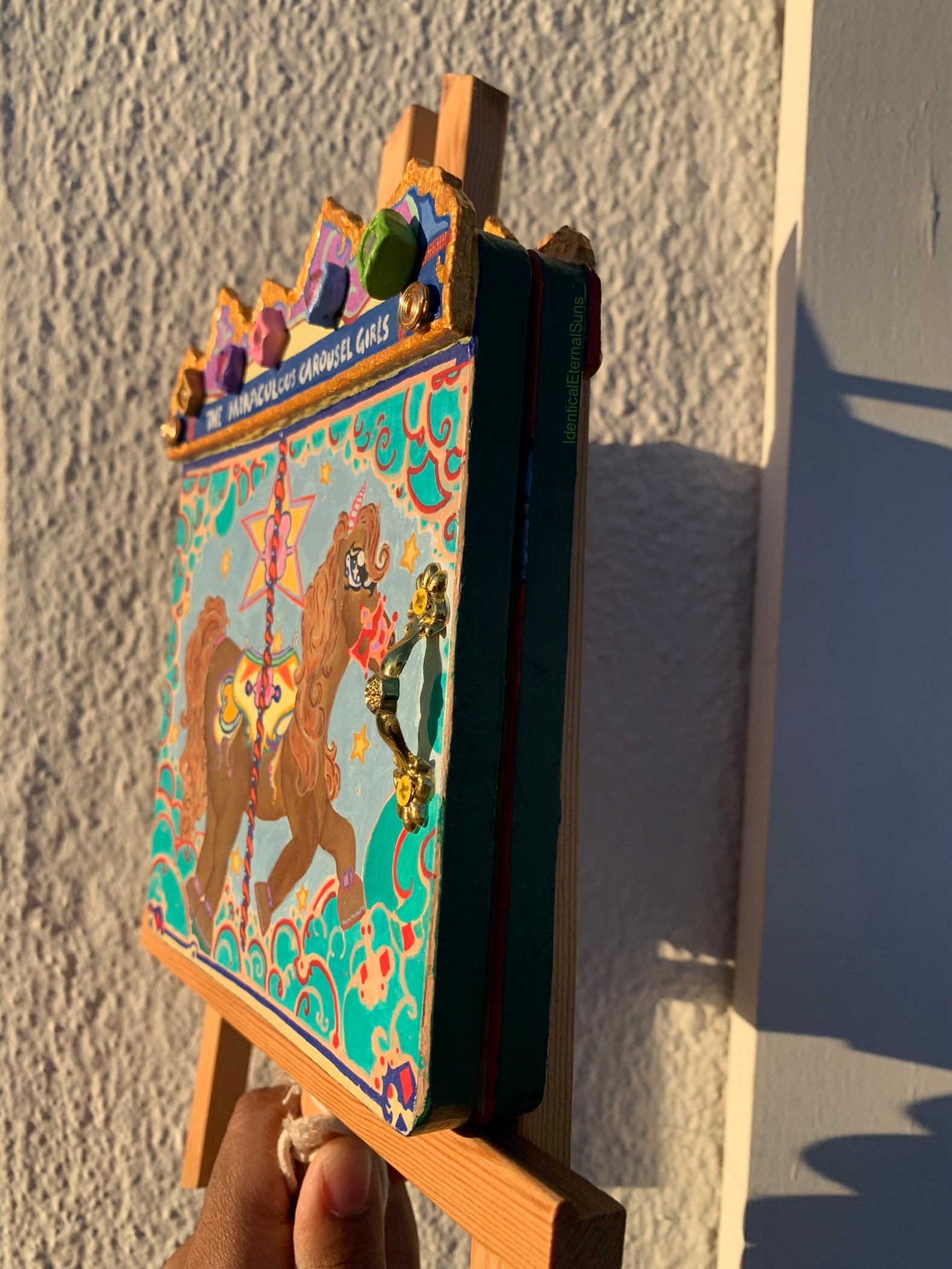 Colorful miniature painting of ponies on a carousel ride on a small easel against a textured wall. Colorful carousel-themed pony mini painting artwork. The Miraculous Carousel Girls wooden painting book, vibrantart, equineart, carouselride, merrygoroundmagic, immersiveart, nostalgickidcore, hopefulart, innocentjoy, nurserydecor, babygirlroom, childlikemagic, playfulimagination, joyfulplaytime, majesticlookbook, nurserybabyart, babygirldecor, cherishablekeepsakes, functionalsurrealism, surrealismsculpture