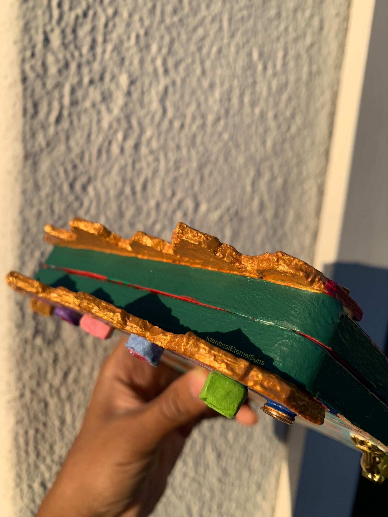 Colorful carousel-themed pony mini painting artwork. The Miraculous Carousel Girls wooden painting book, vibrantart, equineart, carouselride, merrygoroundmagic, immersiveart, nostalgickidcore, hopefulart, innocentjoy, nurserydecor, babygirlroom, childlikemagic, playfulimagination, joyfulplaytime, majesticlookbook, nurserybabyart, babygirldecor, cherishablekeepsakes, functionalsurrealism, surrealismsculpture 
Decorative item with gold and green colors held against a textured wall.