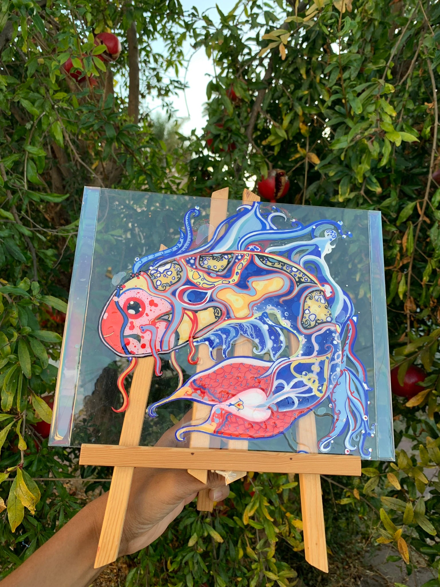 Colorful fish painting on a small easel held in front of green foliage with red fruit. Identicaleternalsuns, art collection, art buyer, Identicaleternalsuns, DenleyTwins, Whimsical, radiant, otherworldly, glorious, fantastical, eternal, divine, Dreamy Fish, Aquarium wonderland, Frutiger Aero vibes, identicaleternalsuns, Ecstatic, frutiger aero art, fish painting on glass, koi, koifish art, mixed media , fishart, fish glass painting, iridescentart, underwaterart, traditional art, contemporary art, asian art