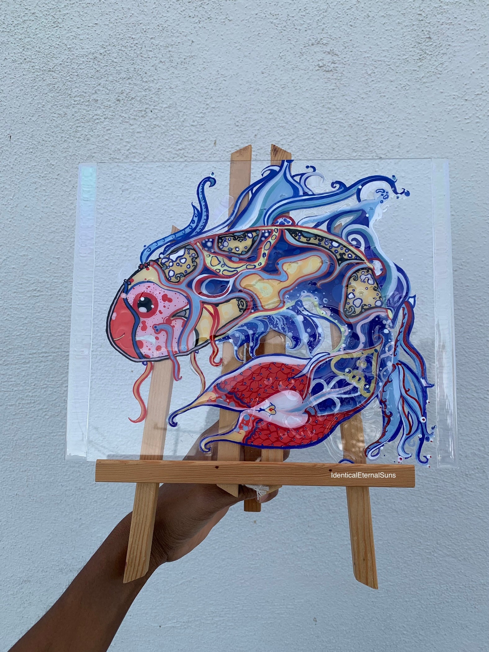 Identicaleternalsuns, art collection, art buyer, Identicaleternalsuns, DenleyTwins, Whimsical, radiant, otherworldly, glorious, fantastical, eternal, divine, Dreamy Fish, Aquarium wonderland, Frutiger Aero vibes, identicaleternalsuns, Ecstatic, frutiger aero art, fish painting on glass, koi, koifish art, mixed media , fishart, fish glass painting, iridescentart, underwater, traditional art Colorful abstract fish drawing on a clear sheet held by a person with a wooden easel against a light gray background.