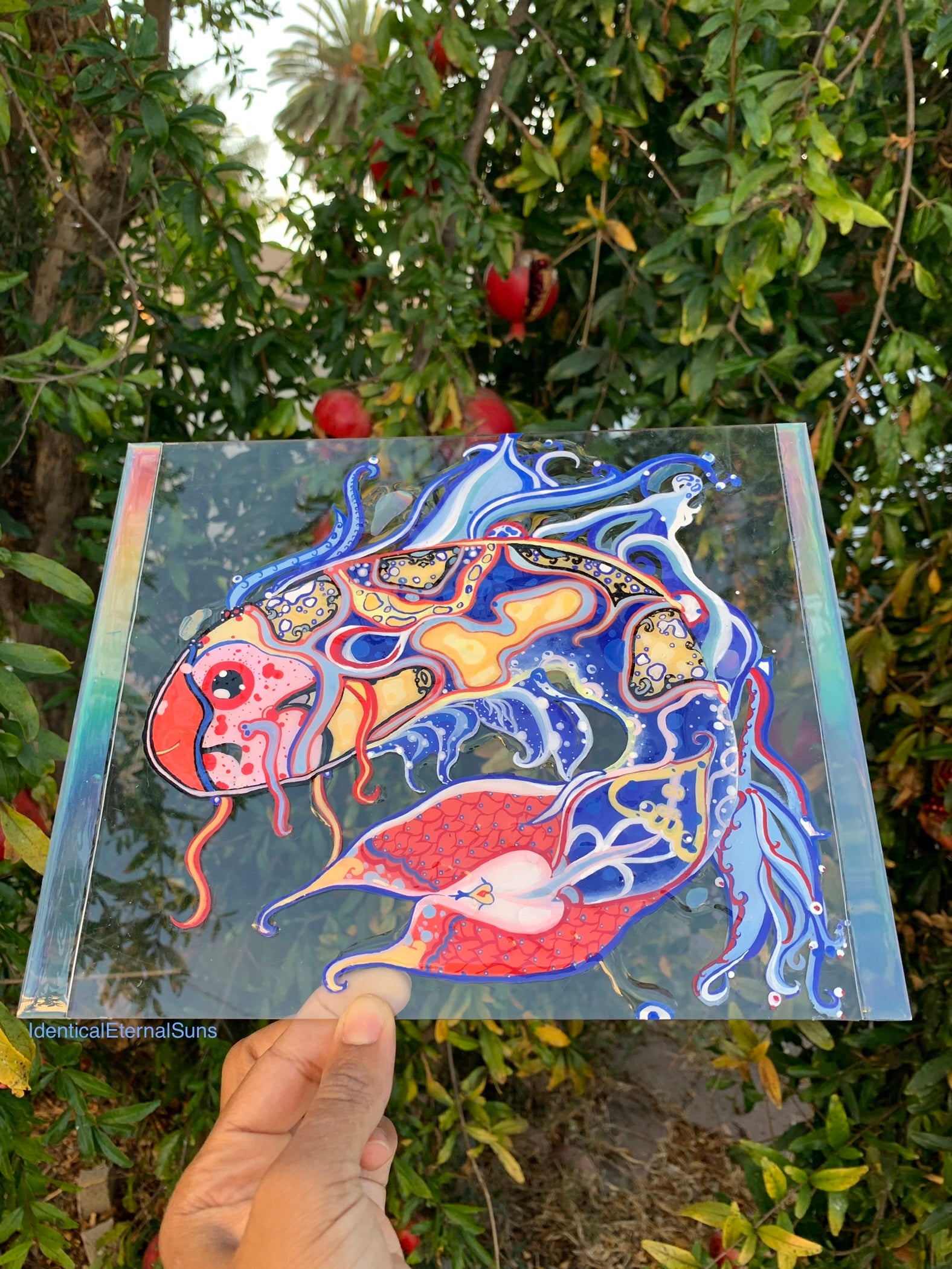 Colorful fish artwork held in front of a tree. Identicaleternalsuns, art collection, art buyer, Identicaleternalsuns, DenleyTwins, Whimsical, radiant, otherworldly, glorious, fantastical, eternal, divine, Dreamy Fish, Aquarium wonderland, Frutiger Aero vibes, identicaleternalsuns, Ecstatic, frutiger aero art, fish painting on glass, koi, koifish art, mixed media , fishart, fish glass painting, iridescentart, underwaterart, traditional art, contemporary art, asian art, nature art, vibrant colors, handmade