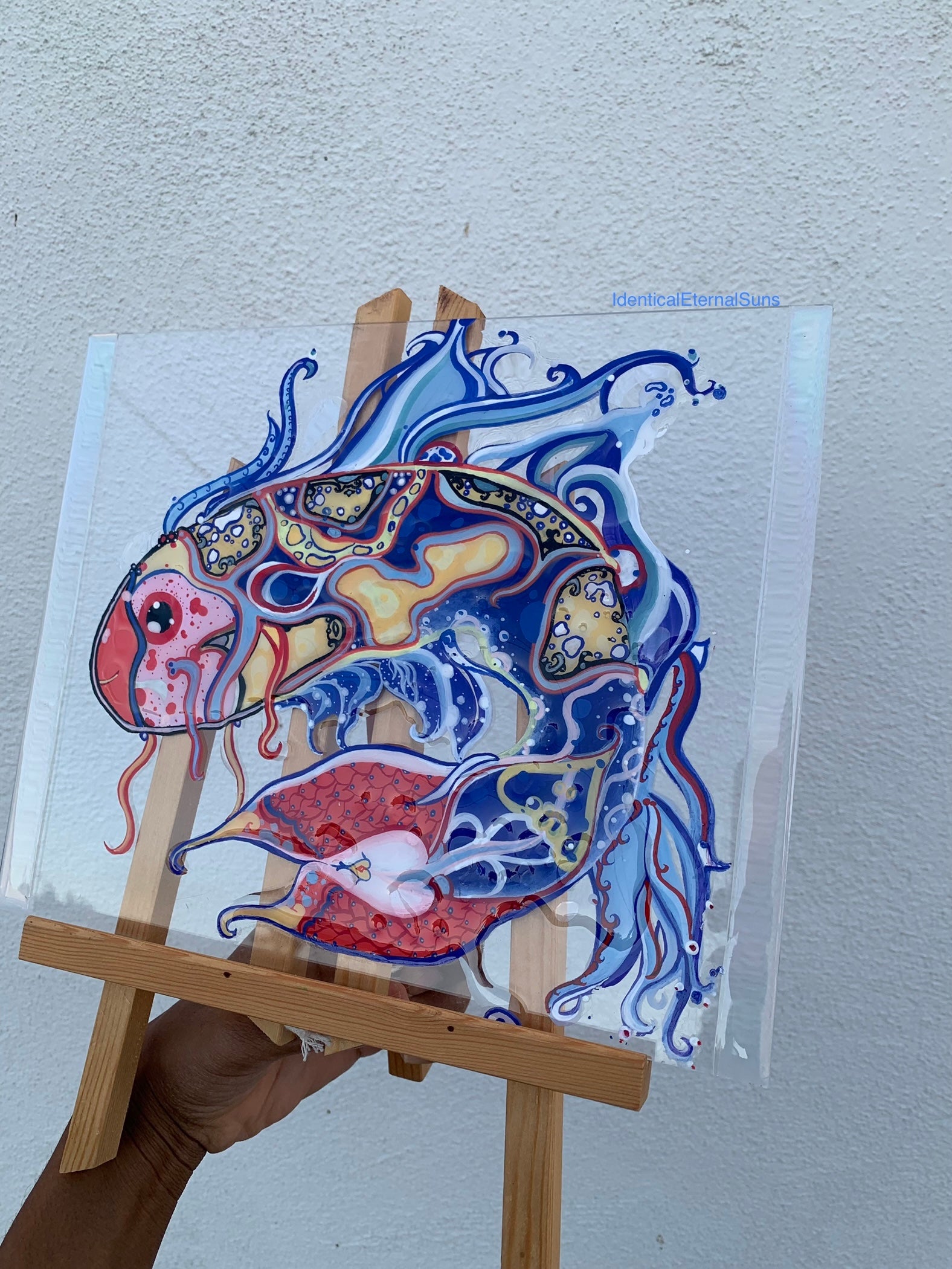 Identicaleternalsuns, art collection, art buyer, Identicaleternalsuns, DenleyTwins, Whimsical, radiant, otherworldly, glorious, fantastical, eternal, divine, Dreamy Fish, Aquarium wonderland, Frutiger Aero vibes, identicaleternalsuns, Ecstatic, frutiger aero art, fish painting on glass, koi, koifish art, mixed media , fishart, fish glass painting, iridescentart, underwaterart, traditional art, Colorful abstract fish painting on a clear sheet held by a person with a wooden easel against a light gray wall.