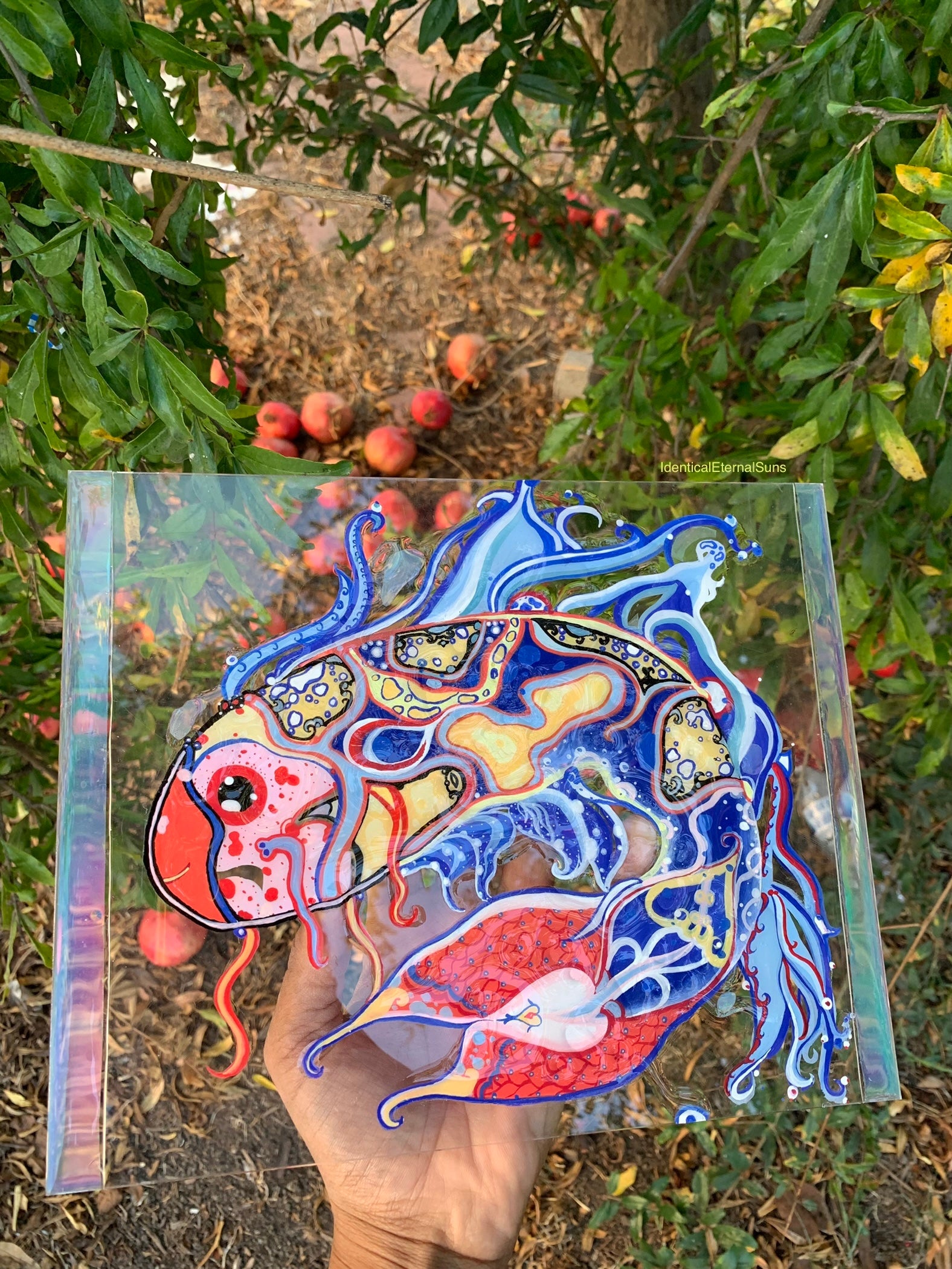 Colorful fish-shaped glass panel held in front of a natural background with trees and fruits. Identicaleternalsuns, art collection, art buyer, Identicaleternalsuns, DenleyTwins, Whimsical, radiant, otherworldly, glorious, fantastical, eternal, divine, Dreamy Fish, Aquarium wonderland, Frutiger Aero vibes, identicaleternalsuns, Ecstatic, frutiger aero art, fish painting on glass, koi, koifish art, mixed media , fishart, fish glass painting, iridescentart, underwaterart, traditional art, contemporary art