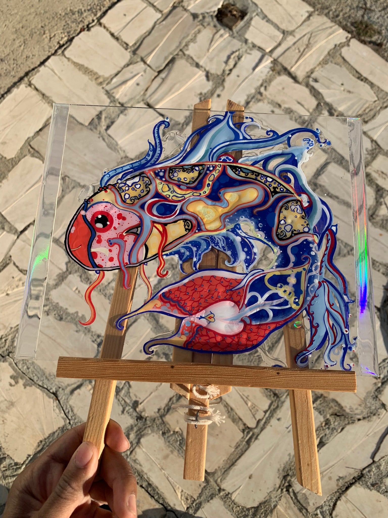 Colorful fish artwork on a transparent board held by a hand on a stone pavement. Identicaleternalsuns, art collection, art buyer, Identicaleternalsuns, DenleyTwins, Whimsical, radiant, otherworldly, glorious, fantastical, eternal, divine, Dreamy Fish, Aquarium wonderland, Frutiger Aero vibes, identicaleternalsuns, Ecstatic, frutiger aero art, fish painting on glass, koi, koifish art, mixed media , fishart, fish glass painting, iridescentart, underwaterart, traditional art, contemporary art, asian art